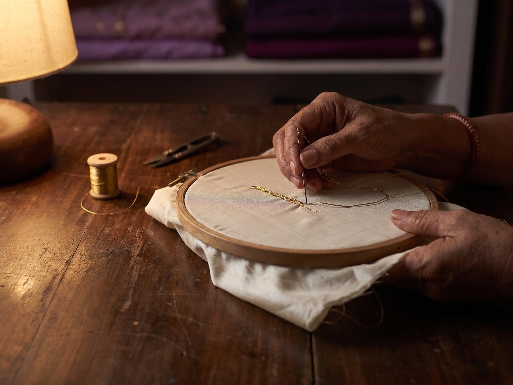 Hands stitching gold zari thread into ivory cloth held in a wooden embroidery frame, under a low lamp