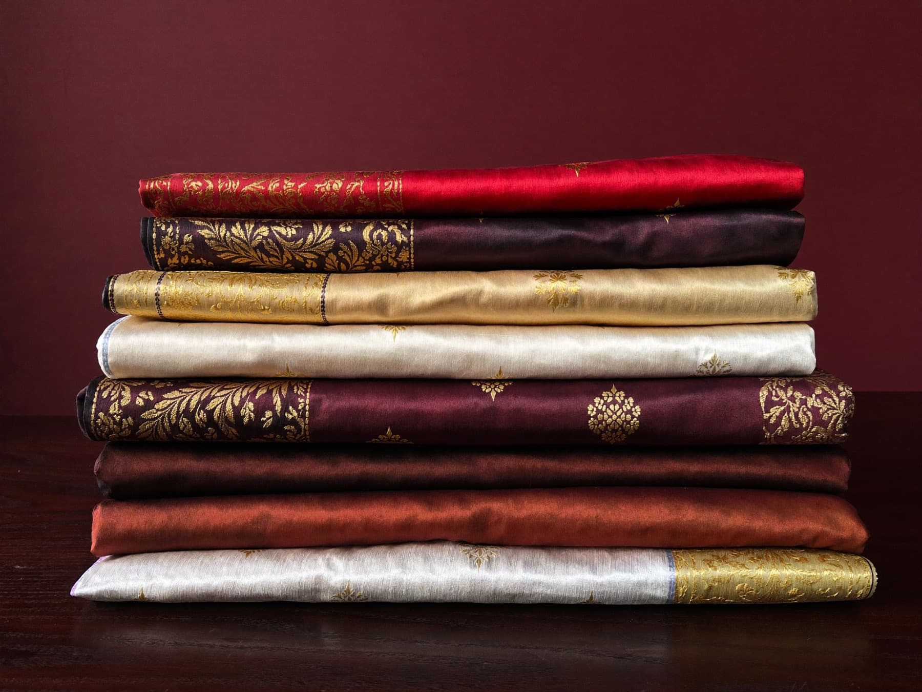 Folded silk cloth in crimson, ivory, indigo and gold stacked on a dark wood table